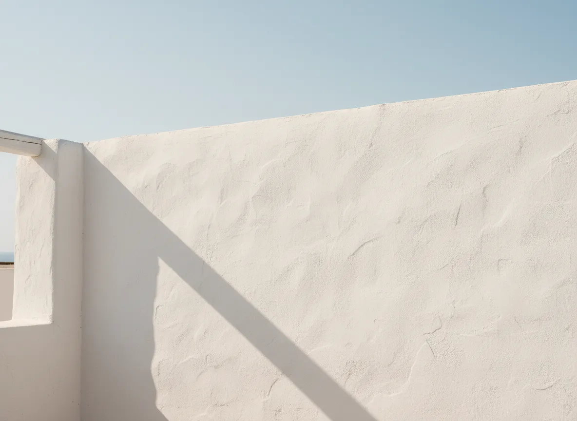 Clean whitewashed stucco exterior wall under open sky, simple and bright with a faint surface texture and a sliver of shadow line where wall meets ground.